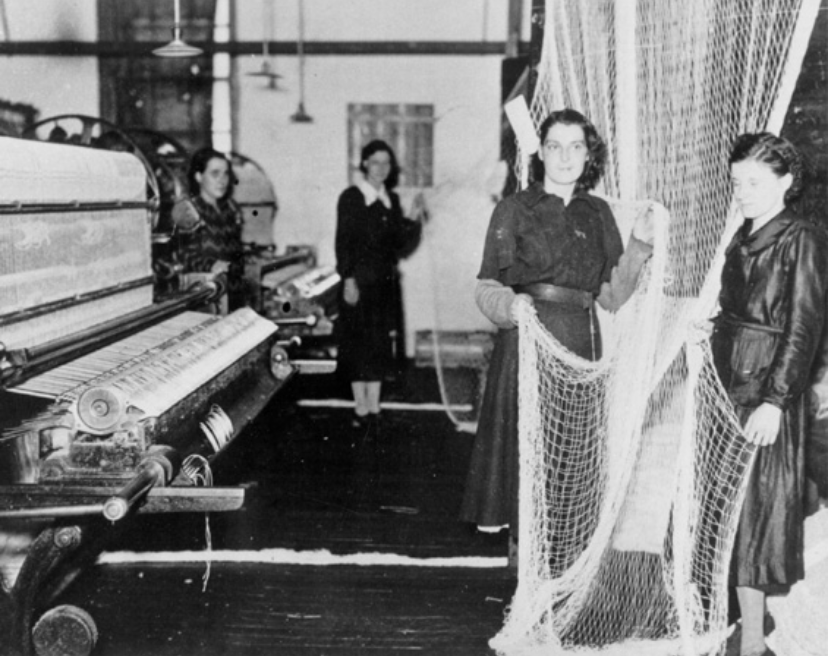 Dans une vieille photo en noir et blanc, deux femmes sont debout à la droite d'une machine de fabrication de filet, elles en tiennent un dans leurs mains. En arrière deux autres femmes sont debout à la gauche.